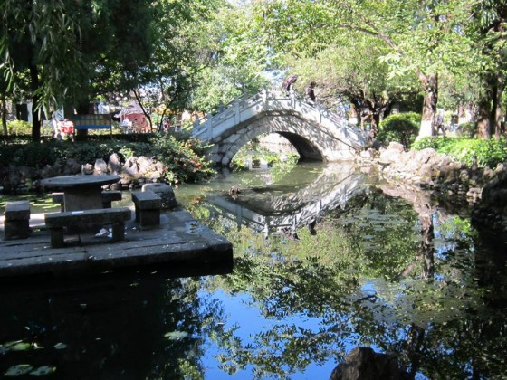 A couple crossing a bridge in a Dali park are reflected in the pond's waters.