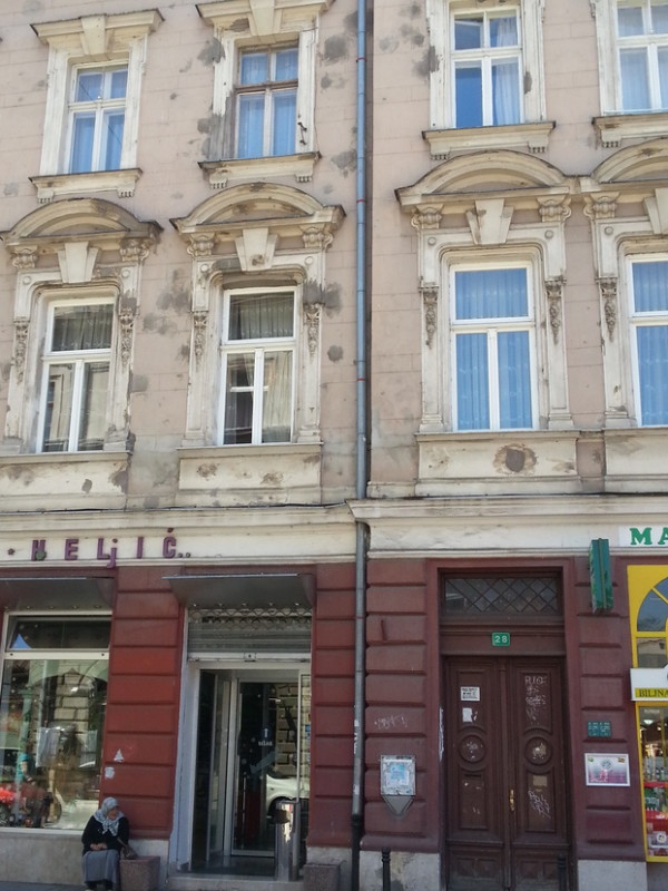 An old woman sits on a stoop begging from passersby; behind her, a building showing repairs from bullets and mortar fire.