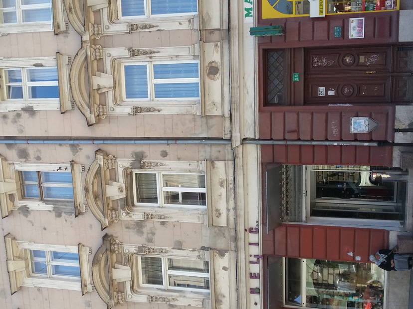An old woman sits on a stoop begging from passersby; behind her, a building showing repairs from bullets and mortar fire.